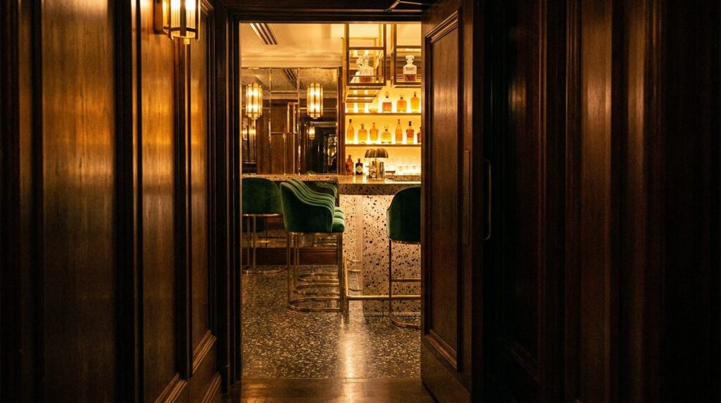 Unmarked wooden door at the end of a dim hotel corridor opening into a moody art-deco cocktail bar