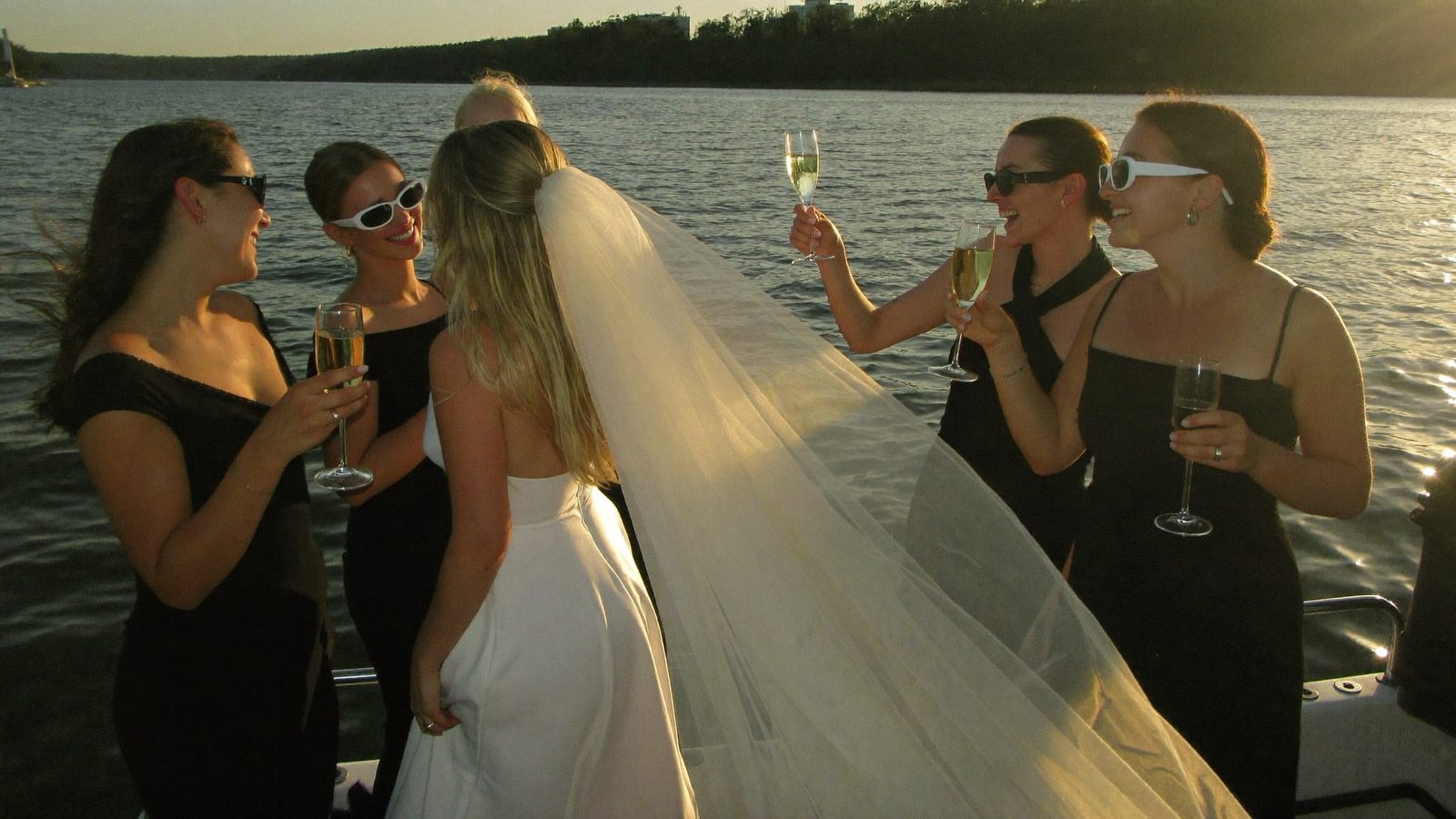 Bride in white veil with crew in black raising a champagne toast at golden hour - Veils and Vows theme